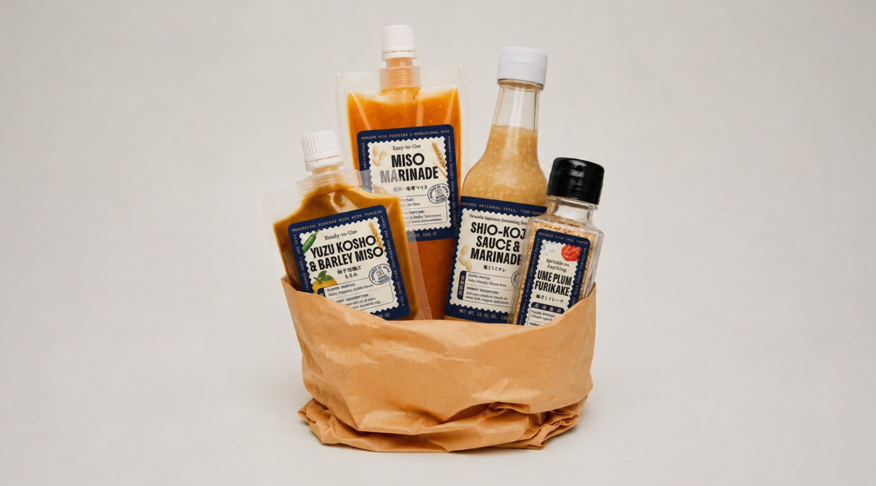 A gift basket of hot sauces and miso marinade in a brown paper bag, photographed on a clean light gray background. The products include Srio-Soji Sriracha & Garlic Marinade, Yuzu Choro & Butter Miso, and Miso Marinade.