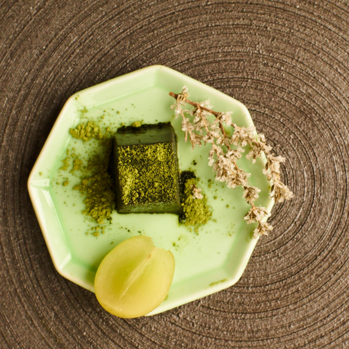A plate of black sesame tofu topped with matcha powder next to sliced lemon